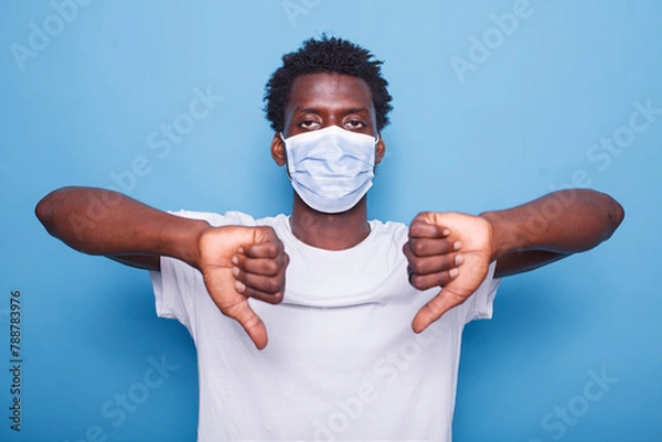 Fototapeta African American adult showing thumbs down sign with both hands. Black man gesturing with his fingers while looking at camera and wearing face mask to protect from coronavirus epidemic.