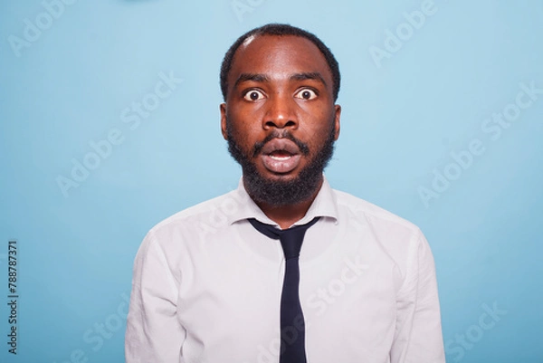 Obraz Stunned businessman stands in front of an unbelievable situation on a blue background, his mouth hanging open in shock. Surprised black individual with wide eyes and a jaw drop.