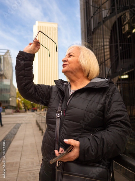 Fototapeta senior blonde business woman looks at her glasses with cell phone in hand in winter in office district