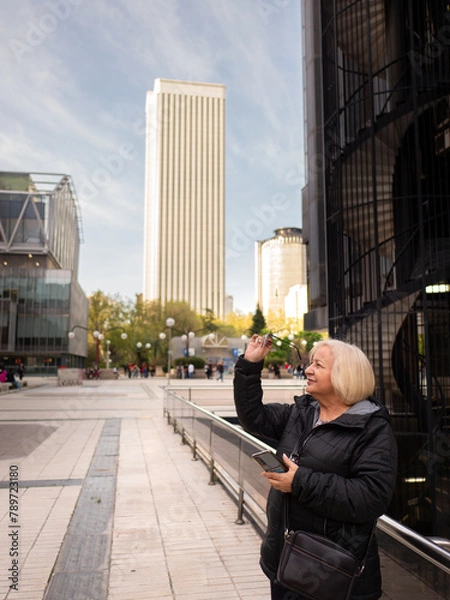 Fototapeta senior blonde business woman looks at her glasses with cell phone in hand in winter in office district