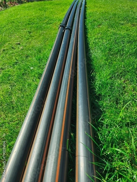 Fototapeta Close-up of four black plastic pipes neatly arranged on lush green grass field under bright sunlight, showcasing contrast between man-made objects and natural environment.