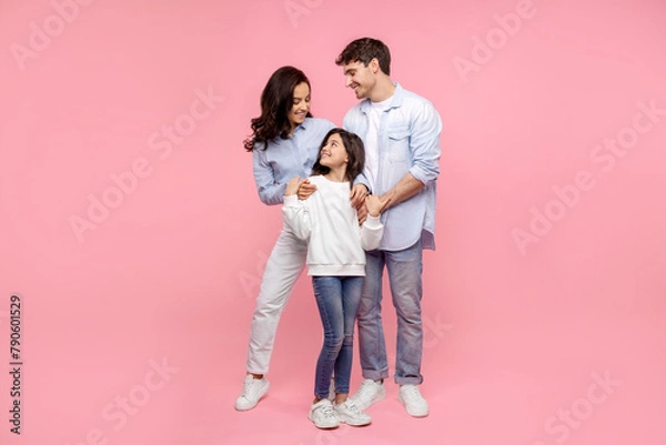 Fototapeta European parents embracing their happy daughter looking at pretty girl, standing together on pink studio background, full length shot