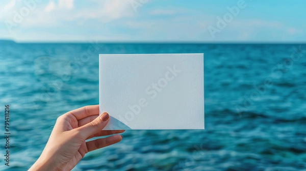 Fototapeta A woman's hand holds a white blank piece of paper against the background of the sea. Mockup, product advertisement, invitation flyer