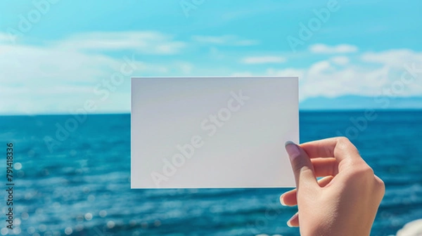 Fototapeta A woman's hand holds a white blank piece of paper against the background of the sea. Mockup, product advertisement, invitation flyer