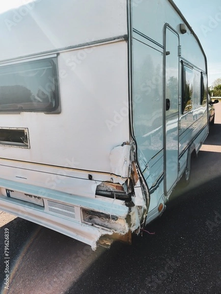 Fototapeta Vintage white travel trailer with a unique retro design and a dent on the back corner, parked by a tranquil lake surrounded by lush green trees, creating a serene setting for a peaceful retreat.