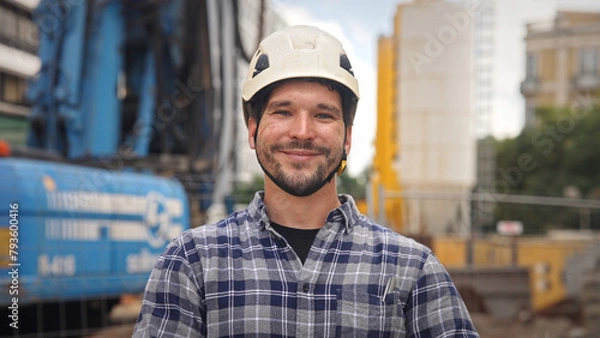 Obraz Confident construction site worker portrait. Smile constructor man look at camera. Builder engineer wear white helmet. Real estate agent plaid shirt. Guy build new house city street Erector person job