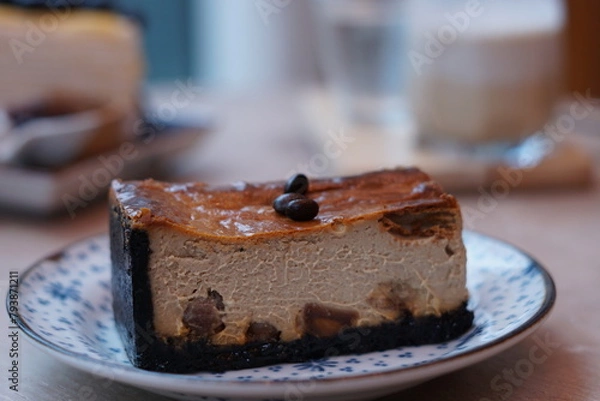 Fototapeta A piece of espresso macadamia cheese cake topped with coffee beans on the plate with blur of blueberry crepe cake and dirty latte coffee.