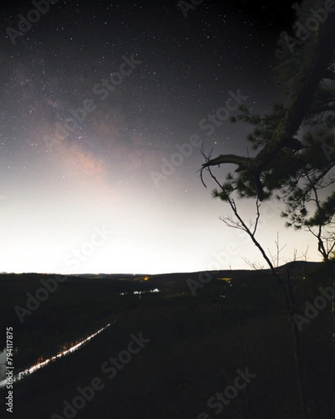Obraz The Milky Way Core above the light dome of the City of Leominster Massachusetts. A road is running towards the milky way with a cars head lights creating a light trail. A lone tree branch points.