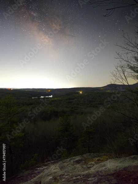 Fototapeta The Milky Way Galaxy as viewed from the top of a small cliff in central Massachusetts. The core of the galaxy is visible. Stones on the top of cliff are in the foreground as well as a large forest.