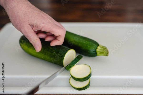Obraz Close-up of older woman's hands slicing zucchini with a knife on a white cutting board horizontal