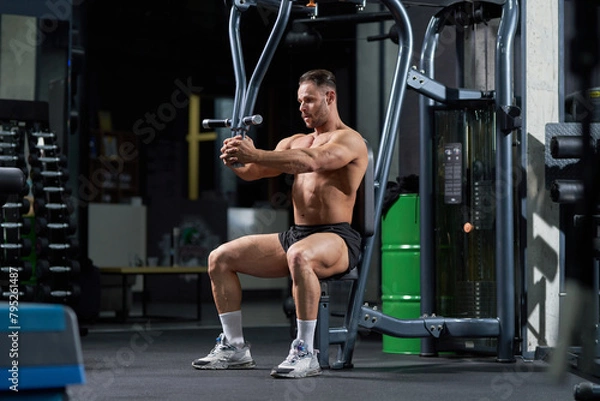Fototapeta Determined middle-aged bodybuilder training at gym, sweating. Side view of attractive Caucasian man using exercising machine during workout in gym. Concept of sporty lifestyle.