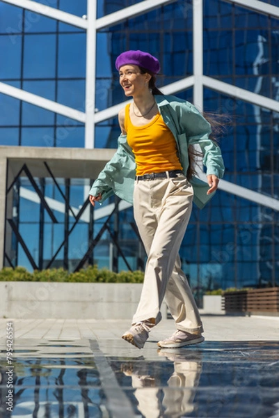 Obraz Young Woman In Stylish Outfit Walking Joyfully In Urban Setting With Glass Building Background, Reflecting Her Movement On Wet Surface. Vertical Screen, Perfect For Lifestyle And Fashion Content.