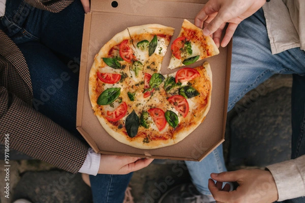 Fototapeta Top view image of couple grab slices of pizza from box at the outdoor. Man and woman hands taking pizza. Vegan pizza with fresh tomatoes basil and broccoli. Lactose and gluten free