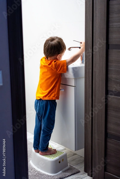 Fototapeta A young boy is standing on a stool in front of a sink. He is washing his hands