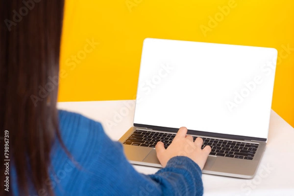 Fototapeta Woman Working on Laptop with Blank Screen,Rear view of a woman using a laptop with a mock-up screen against a vibrant yellow background.
