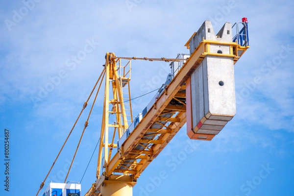 Fototapeta Tower Crane Against Blue Sky: Close-up view of a towering yellow crane with a heavy concrete counterweight under a clear blue sky