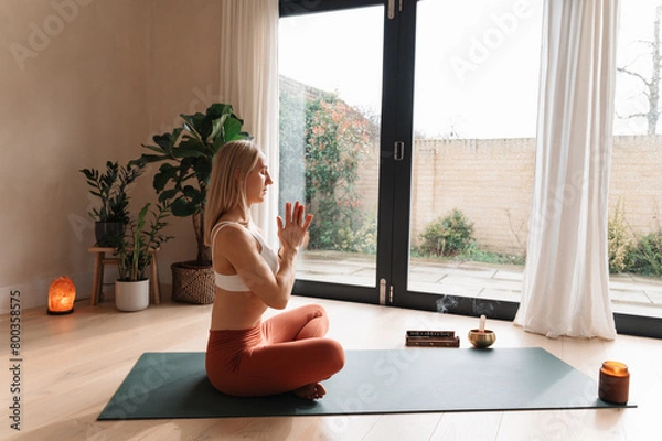 Fototapeta Young woman sitting in lotus pose on yoga mat, holding hands to heart, meditating at home. Copy space. 