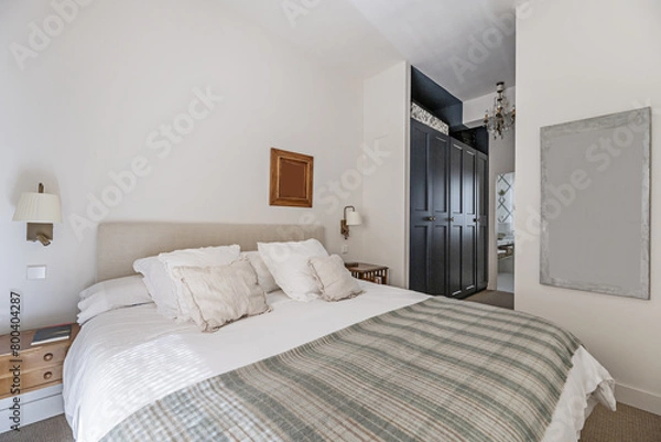 Fototapeta Double bedroom with fabric upholstered headboard and many cushions and pillows on the mattress, a hallway with a walk-in wardrobe and access to a bathroom