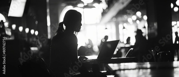 Obraz Silhouette of a woman working on a laptop in a cafe with low light. A black and white photo with a blurred background of people sitting at tables. 