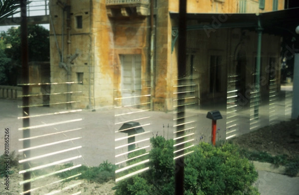 Fototapeta horizontal image of an old yellow building reflected in a glass window. patterns showing horizontal blinds. sunlight and green plants. location Birchircara Rail Station Malta 2003