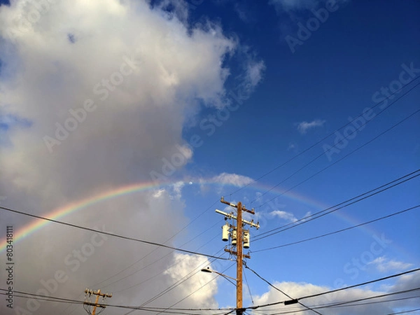 Fototapeta Vibrant Rainbow Peeks Through Clouds Above Power Lines On A Clear Day