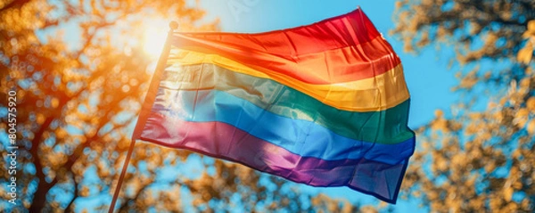 Fototapeta A rainbow flag is flying in the sky. The flag is colorful and vibrant, and it is a symbol of pride and acceptance. The flag is held up by a pole, and it is against a backdrop of trees and a blue sky