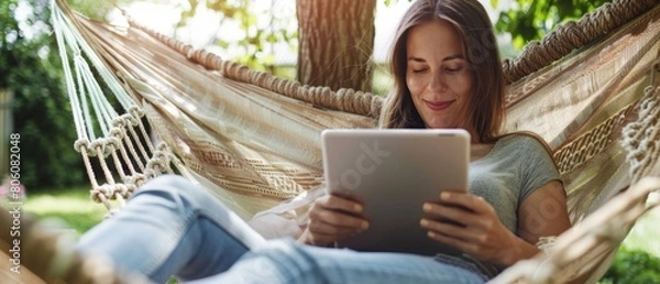 Fototapeta Smiling woman reading on tablet while relaxing in hammock.
