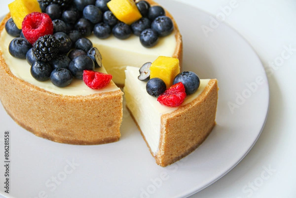 Obraz Cheesecake decorated with fresh berries and mango. On a light gray plate. White background. Back view. There is a copy space for your text.