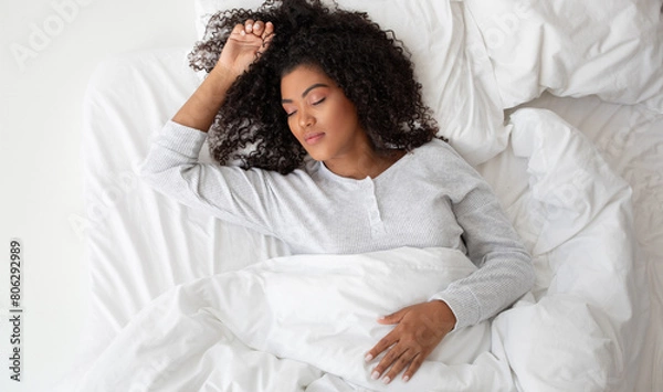 Fototapeta A Hispanic woman is laying comfortably in a bed covered with crisp white sheets. She appears relaxed and is surrounded by a serene atmosphere.
