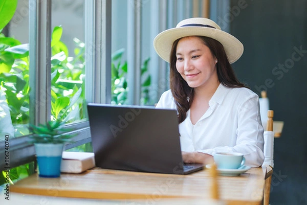 Fototapeta Portrait image of a young woman with hat working on laptop computer in cafe