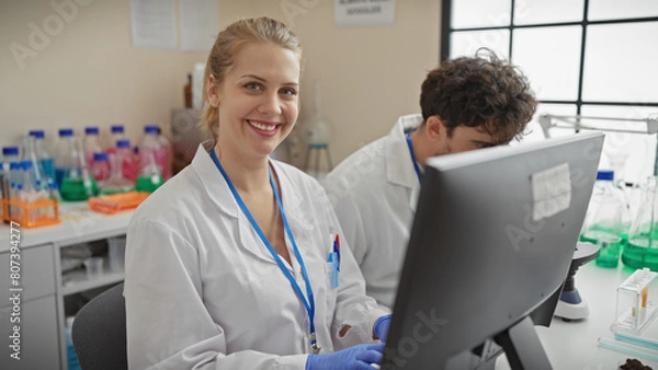 Fototapeta A smiling woman and focused man in white lab coats work together in a brightly lit laboratory setting, surrounded by scientific equipment.