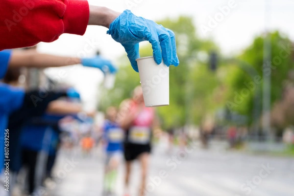 Fototapeta Runners of a marathon are provided with water at a hydration station. Running event, refreshment point along the route of long-distance run.