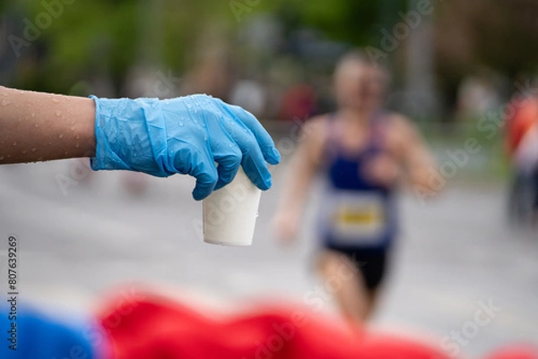 Fototapeta Runners of a marathon are provided with water at a hydration station. Running event, refreshment point along the route of long-distance run.