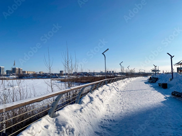 Obraz Une promenade enneigée le long d'une rivière gelée avec un pont et des roseaux, des lampadaires le long de la promenade, avec au loin la vue d'une ville
