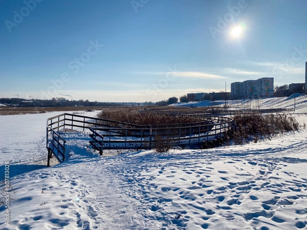 Obraz Une promenade enneigée le long d'une rivière gelée avec un pont et des roseaux, avec au loin la vue d'une ville sous le soleil
