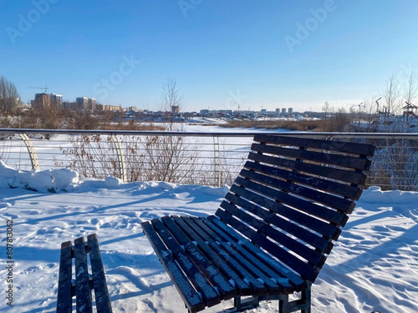 Obraz Une banc sur une promenade enneigée le long d'une rivière gelée avec des roseaux, et au loin, on distingue la silhouette d'une ville