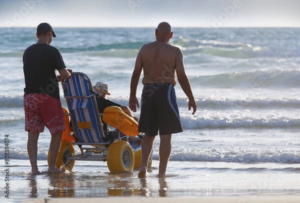 Fototapeta Two men are assisting a elderly man in a specialized beach wheelchair across the sandy shores. Sons taking care of their elderly parent with disabilities, spending time with them by the sea. Care