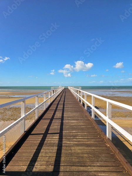 Fototapeta A long wooden pier stretching to the horizon, over a calm beach at low tide with clear blue sky, yellow sand and little white clouds.