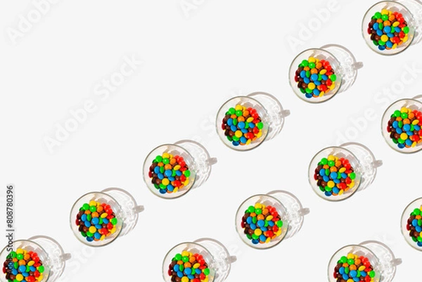 Fototapeta Pattern made of drinking glass full of multicolored candies on a white background with copy space.
