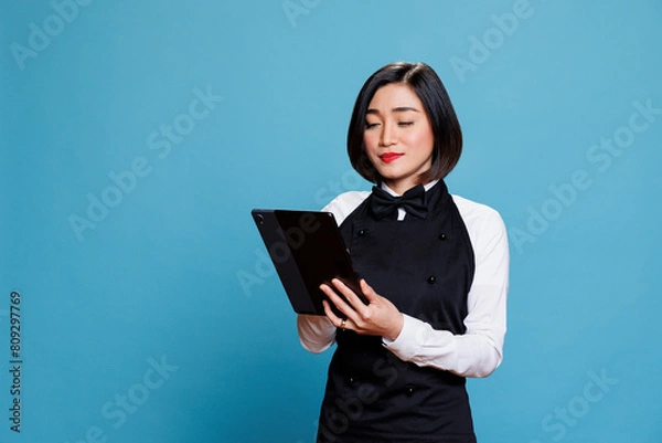 Fototapeta Restaurant young waitress using digital tablet while managing customer order online. Attractive hotel woman receptionist in uniform tapping on portable device touchscreen