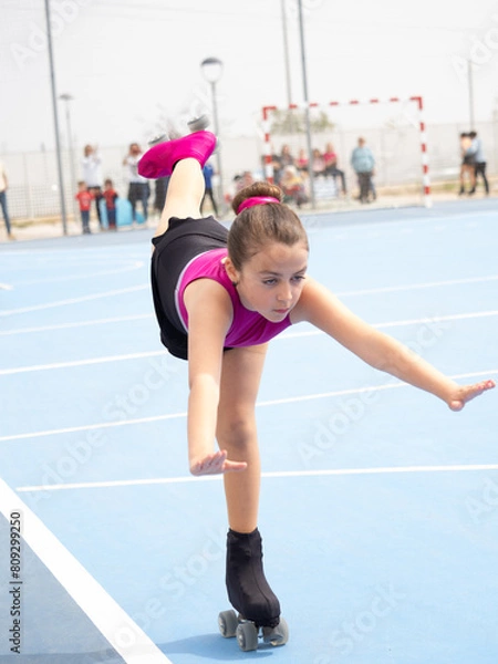 Obraz A young girl in a pink and black outfit performs an artistic roller skating maneuver on an outdoor court.  