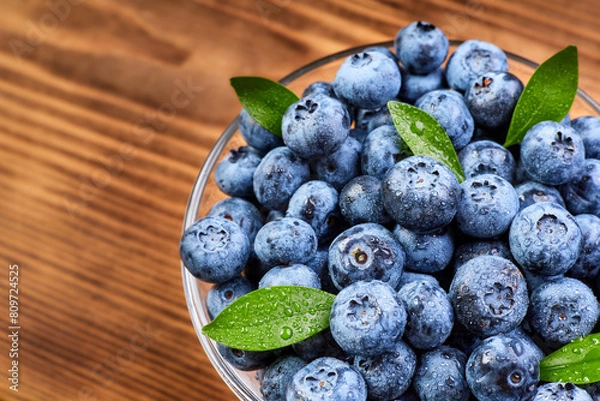 Obraz Blueberries with green leaves in a glass plate and water drops on a wooden background