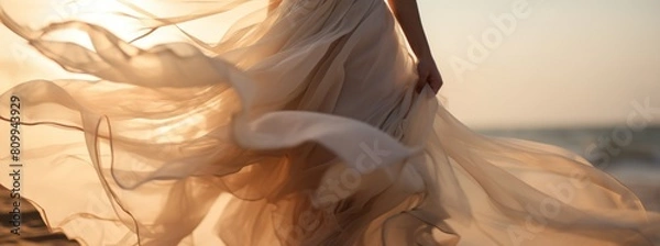 Fototapeta A close-up view captures a woman in a light beige dress billowing in the wind, creating a sense of movement and freedom. There're a sea or the ocean with sand dune on the background.