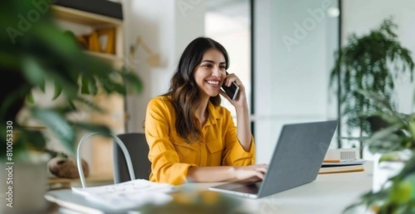 Fototapeta A woman is talking on her cell phone while sitting at a desk with a laptop. She is smiling and she is enjoying her conversation,wearing yellow clothes