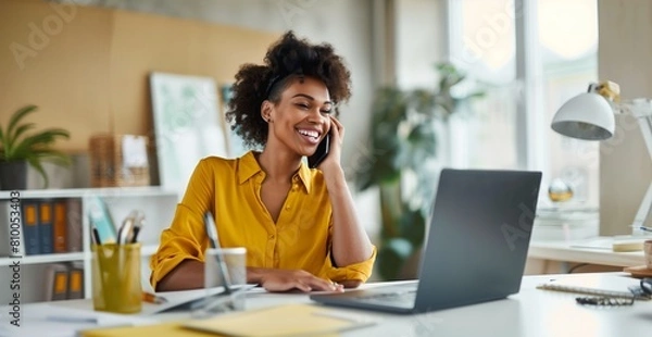 Fototapeta A woman is sitting at a desk with a laptop and a cell phone. She is smiling and talking on the phone,wearing yellow clothes