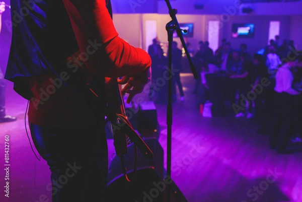 Fototapeta Concert view of an electric guitar player with vocalist and rock band performing in a club, male musician guitarist on stage with audience in a crowded concert hall arena, hands on a bass guitar