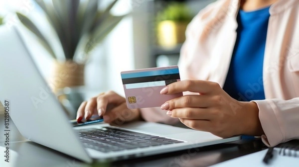 Fototapeta A woman in a pink blazer is holding a credit card and using a laptop. She is shopping online and is about to make a purchase.