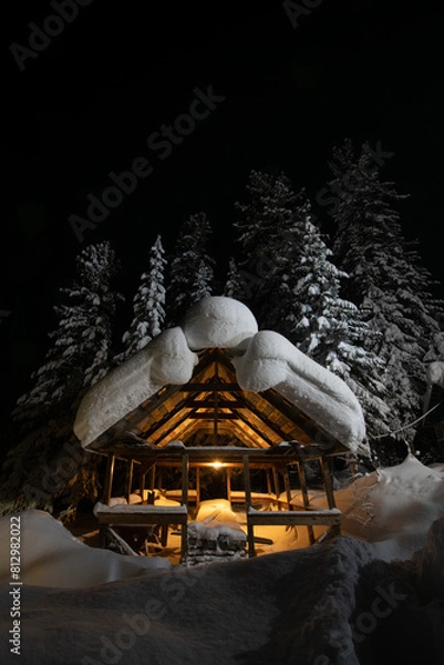 Fototapeta The frame of a house under construction in a Siberian forest illuminated by a lamp in the night with a huge mass of snow lying on the roof