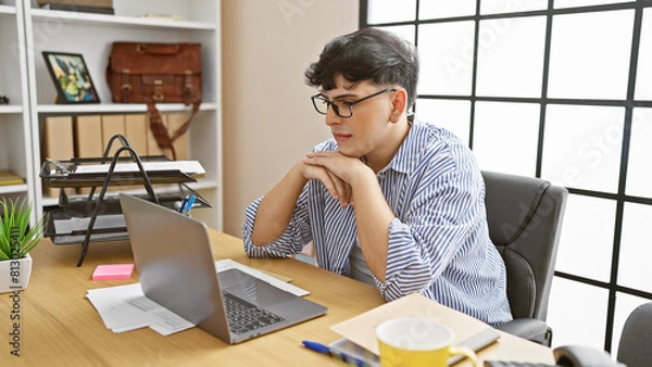 Fototapeta A thoughtful young man with glasses sits at a wooden desk, working on a laptop in a well-lit office.