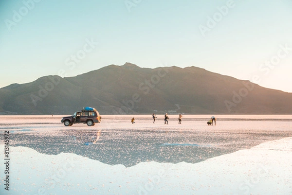 Fototapeta Reflections at Salar de Uyuni during sunset -April- - 4x4 Jeep and tourists behind mountain and mirror lake